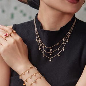 Puede incluir: Juego de joyas doradas con un collar en capas, una pulsera, pendientes y anillos. El collar y la pulsera tienen colgantes colgantes, incluyendo el número ocho y colgantes en forma de estrella. Los anillos tienen piedras preciosas rojas y marrones.