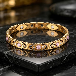 May include: A gold-toned link bracelet featuring an alternating pattern of X-shaped links and oval settings. The oval settings contain round, light pink faceted stones flanked by smaller clear stones. The bracelet includes a secure metal clasp closure.