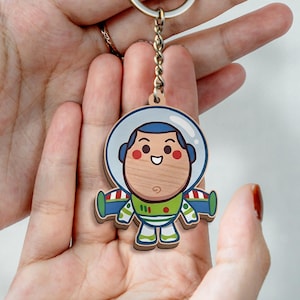 May include: A wooden keychain featuring a cartoon astronaut character with a round face, a white and blue helmet, and a green, white, and purple space suit. The character has a smiling expression, red cheeks, and is attached to a silver metal ring.