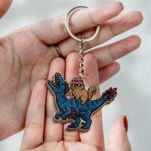 Pode incluir: Um porta-chaves de madeira com um dinossauro azul com detalhes vermelhos, carregando um gato com óculos de sol e uma coroa. O porta-chaves tem um anel e uma corrente prateados.
