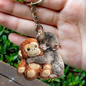 May include: A keychain featuring two monkey figures. One is a plush, orange monkey hugging a realistic, gray monkey. The keychain has a metal ring and chain. The background is a blurred view of green foliage.