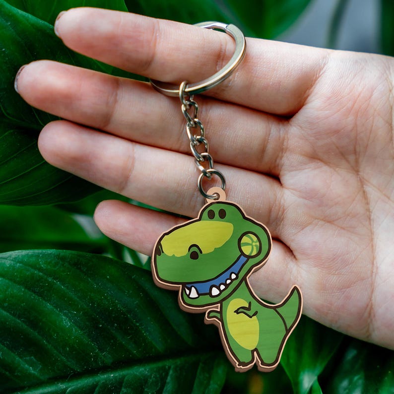 May include: A keychain featuring a cartoon-style green tyrannosaurus rex character with a yellow belly and a small basketball held in its mouth. The dinosaur is attached to a silver-toned metal split ring by a short metal chain link connector.