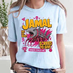 May include: Light blue t-shirt with a graphic featuring a shark-spider hybrid with the name "JAMAL" in yellow block letters. Text includes "HAS SO MANY SORRIES TO GIVE" and "DUNGEON CRAWLER CARL".