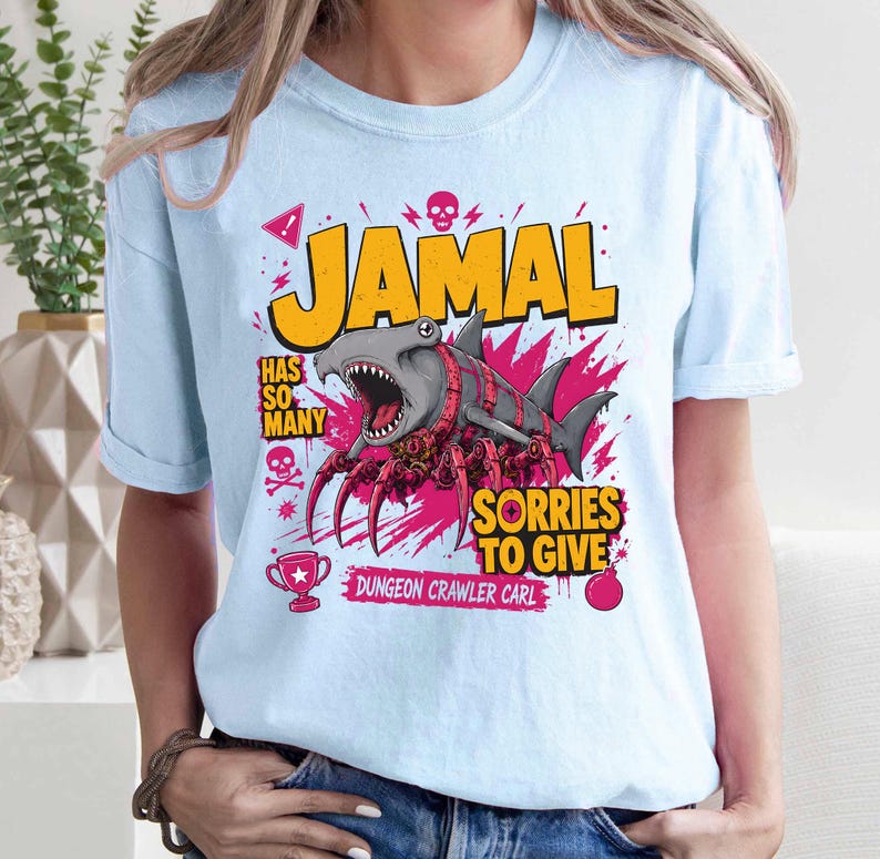 May include: Light blue t-shirt with a graphic featuring a shark-spider hybrid with the name "JAMAL" in yellow block letters. Text includes "HAS SO MANY SORRIES TO GIVE" and "DUNGEON CRAWLER CARL".