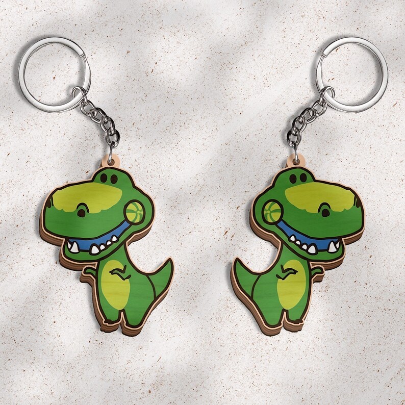 May include: Two identical keychains featuring a cartoon Tyrannosaurus Rex dinosaur in shades of green. The dinosaur has a light green belly, a blue mouth, white teeth, and a small basketball graphic on its cheek, attached to a silver metal ring.