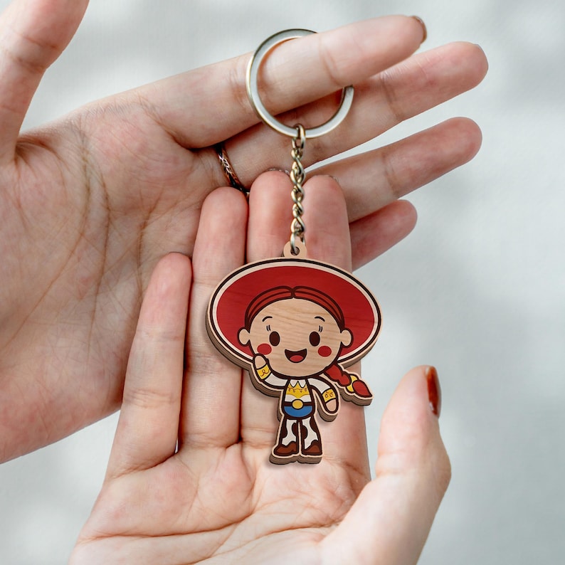 K&ouml;nnte beinhalten: Ein Holzschl&uuml;sselanh&auml;nger mit einer Zeichentrickfigur, die einen gro&szlig;en roten Hut, rotes geflochtenes Haar und ein Cowgirl-Outfit tr&auml;gt. Die Figur hat einen l&auml;chelnden Ausdruck, rote Wangen und ist mit einer kleinen Kette an einem silbernen Ring befestigt.