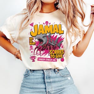 May include: Cream-colored t-shirt with a graphic featuring the name "JAMAL" in yellow, a cartoon shark with pink accents, and the phrase "SORRIES TO GIVE." Additional text includes "HAS SO MANY" and "DUNGEON CRAWLER CARL."