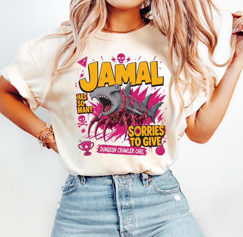 May include: Cream-colored t-shirt with a graphic featuring the name "JAMAL" in yellow, a cartoon shark with pink accents, and the phrase "SORRIES TO GIVE." Additional text includes "HAS SO MANY" and "DUNGEON CRAWLER CARL."