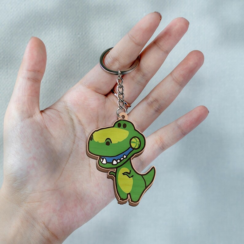May include: A wooden keychain featuring a cartoon-style green dinosaur with a yellow belly and a small ball near its mouth. The dinosaur is attached to a silver-colored metal split ring by a short chain, showcasing a flat, laser-cut design.