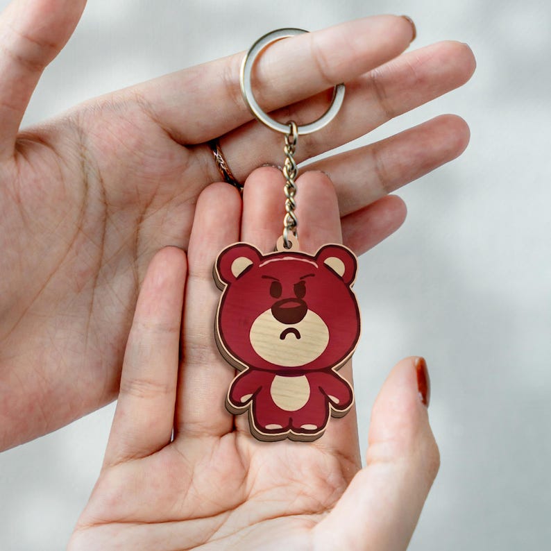 May include: A wooden keychain featuring a cartoon bear with a deep red body, a light tan face, and a dark brown nose. The bear has a grumpy facial expression with furrowed eyebrows and a curved mouth. It is attached to a silver metal ring.