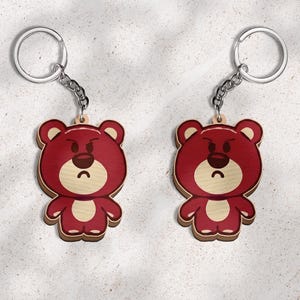 May include: Two identical keychains featuring a flat, wooden bear character with a deep red body, a light beige muzzle, and a dark brown nose. The bear has a grumpy facial expression with furrowed eyebrows and a downturned mouth, attached to a silver metal ring.