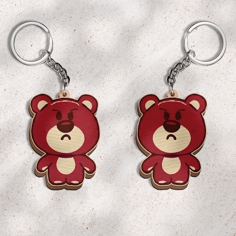 May include: Two identical keychains featuring a flat, wooden bear character with a deep red body, a light beige muzzle, and a dark brown nose. The bear has a grumpy facial expression with furrowed eyebrows and a downturned mouth, attached to a silver metal ring.