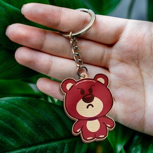 May include: A wooden keychain featuring a cartoon bear with a dark red body, a light tan face, and a light tan belly. The bear has a grumpy facial expression with furrowed eyebrows and a curved mouth. It is attached to a silver metal ring.