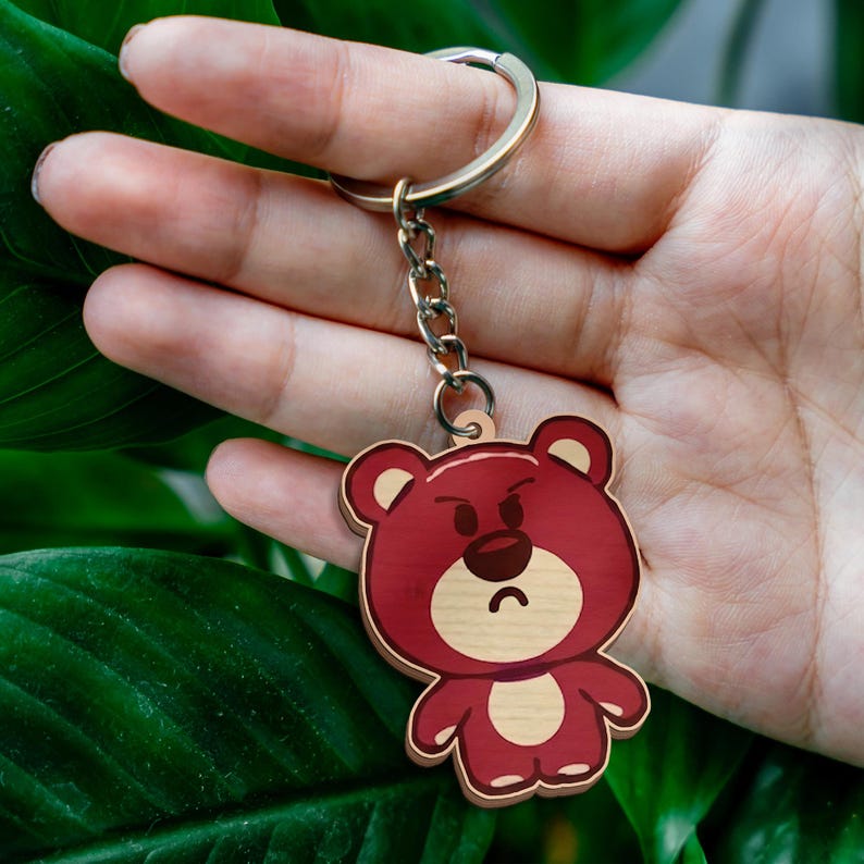 May include: A wooden keychain featuring a cartoon bear with a dark red body, a light tan face, and a light tan belly. The bear has a grumpy facial expression with furrowed eyebrows and a curved mouth. It is attached to a silver metal ring.