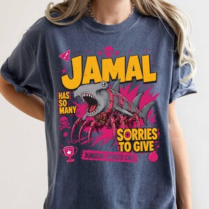 May include: A navy blue t-shirt with a graphic design featuring the name "JAMAL" in yellow block letters. The design includes a shark with spider-like legs, the text "HAS SO MANY" and "SORRIES TO GIVE", plus a trophy and skull icons.