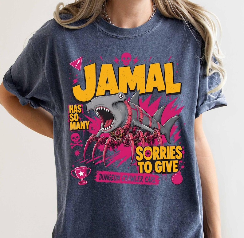 May include: A navy blue t-shirt with a graphic design featuring the name "JAMAL" in yellow block letters. The design includes a shark with spider-like legs, the text "HAS SO MANY" and "SORRIES TO GIVE", plus a trophy and skull icons.