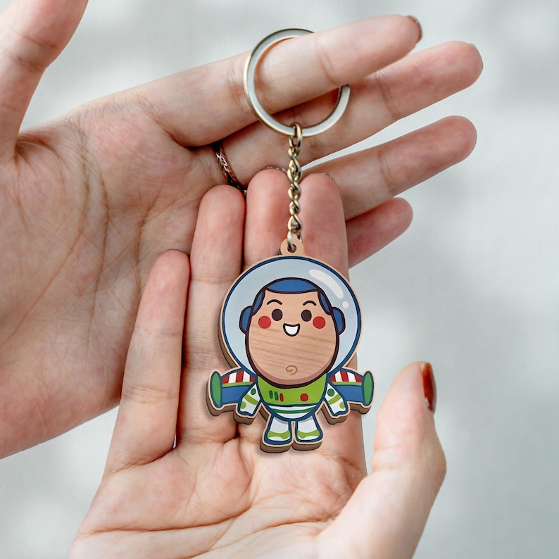 May include: A wooden keychain featuring a cartoon astronaut character with a round face, a white and blue helmet, and a green, white, and purple space suit. The character has a smiling expression, red cheeks, and is attached to a silver metal ring.