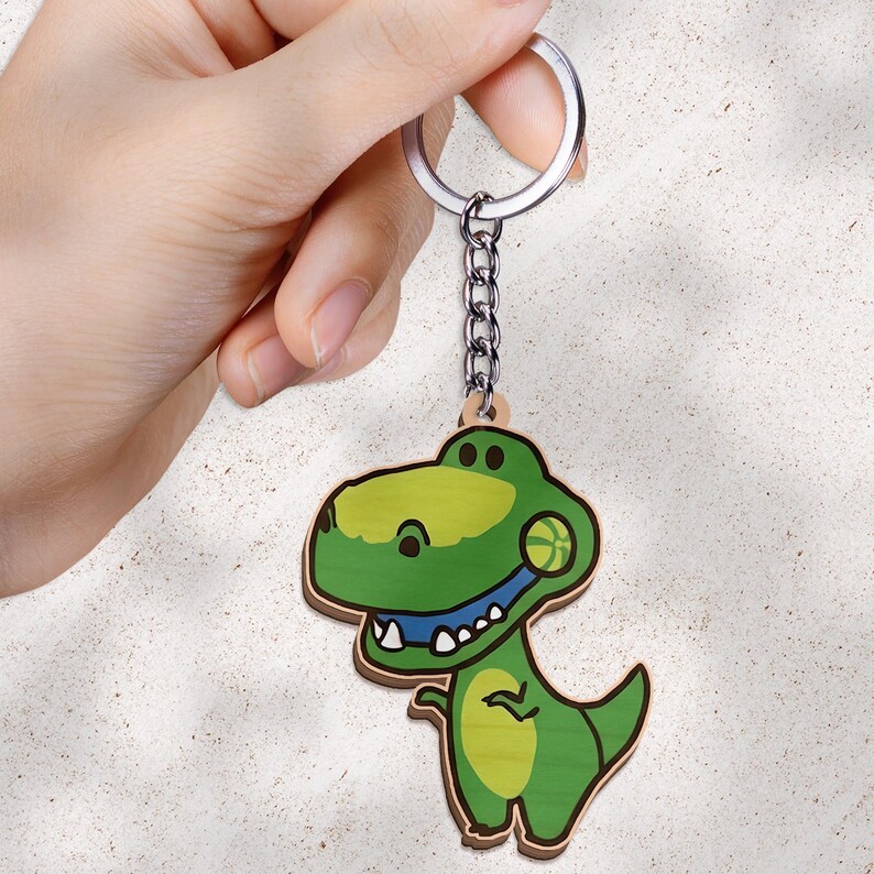 May include: A keychain featuring a flat, cartoon-style green Tyrannosaurus Rex dinosaur holding a small yellow basketball in its mouth. The dinosaur has a light green belly, dark green body, and white teeth. It is attached to a silver metal ring.