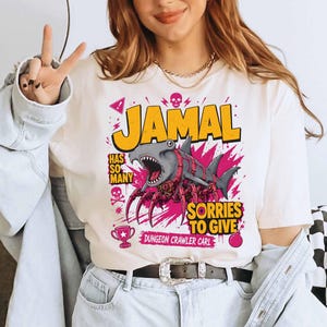 May include: Cream-colored t-shirt with a graphic featuring the name "JAMAL" in yellow, a shark-spider hybrid, and the text "HAS SO MANY SORRIES TO GIVE". Additional graphics include a skull, a trophy, and the words "DUNGEON CRAWLER CARL".