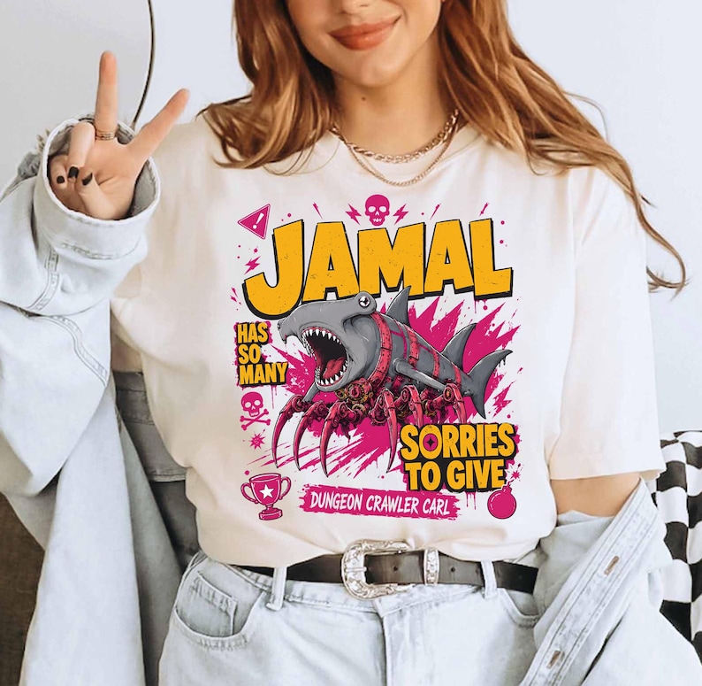 May include: Cream-colored t-shirt with a graphic featuring the name "JAMAL" in yellow, a shark-spider hybrid, and the text "HAS SO MANY SORRIES TO GIVE". Additional graphics include a skull, a trophy, and the words "DUNGEON CRAWLER CARL".