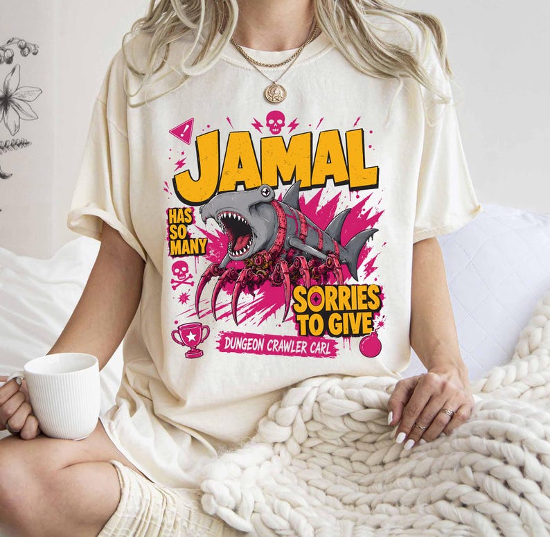 May include: Cream-colored t-shirt featuring a graphic of a shark with spider-like legs and the name "JAMAL" in yellow. The design includes the text "HAS SO MANY SORRIES TO GIVE" and "DUNGEON CRAWLER CARL".