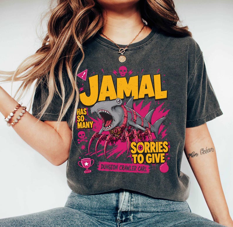 May include: Dark gray t-shirt with a graphic design featuring the name "JAMAL" in yellow, a cartoon shark, and the text "HAS SO MANY SORRIES TO GIVE". The design includes pink and gray accents, skulls, and a trophy.