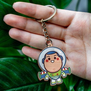 May include: A wooden keychain featuring a cartoon space ranger character with a white helmet, blue hood, green chest plate, and purple wings. The character has a light wood grain face, red cheeks, and a smiling expression, attached to a metal ring.