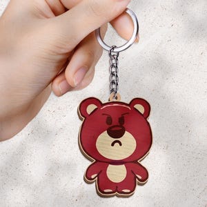 May include: A wooden keychain featuring a cartoon bear with a deep red body, a light beige muzzle, and a matching beige belly patch. The bear has a grumpy facial expression with furrowed brows and a downturned mouth. It is attached to a silver metal ring.