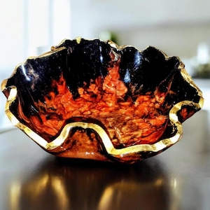 May include: A decorative bowl with a unique, organic shape. The interior features a fiery blend of orange and red hues, contrasted by a glossy black rim. A gold-colored trim outlines the bowl's edge.