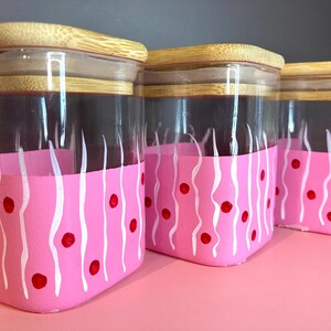 Op de afbeelding: Drie glazen potten met bamboe deksels. Het onderste deel van elke pot is roze geverfd met een wit golvend patroon en rode stippen. De potten staan op een roze ondergrond.