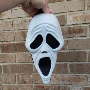 Op de afbeelding: Een wit Scream-masker met zwarte oog- en mondopeningen. Het masker heeft een verweerde, getextureerde uitstraling en wordt tegen een bakstenen muur gehouden. Het ontwerp van het masker heeft een wijd open mond en een bedroefde uitdrukking.