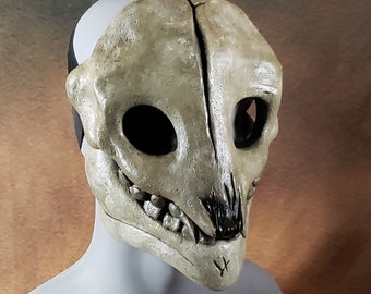 May include: A beige and black animal skull mask with a toothy grin. The mask is made of a smooth, sculpted material and has a realistic appearance.
