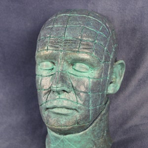 Może przedstawiać: Zielony, fakturowany gipsowy popiersie ludzkiej głowy z siatką na twarzy. Popiersie jest rzeźbą ludzkiej głowy.