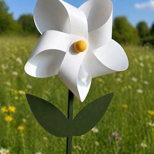 Könnte beinhalten: Eine weiße Papierblume mit gelber Mitte, grünem Stiel und Blättern. Die Blume wird vor einem Feld mit Wildblumen unter blauem Himmel gehalten.