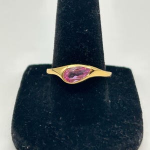 May include: A gold ring featuring a pear-shaped, pink gemstone. The gemstone is set in a gold band with a smooth, polished finish. The ring is displayed on a black velvet ring holder against a white background.