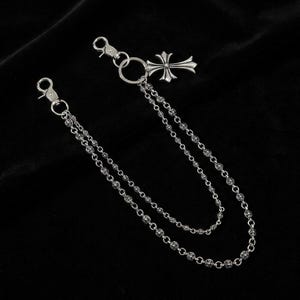 May include: A silver-toned wallet chain with two strands of small, decorative beads. The chain features a cross pendant and two clasps for attachment. The background is a solid black.