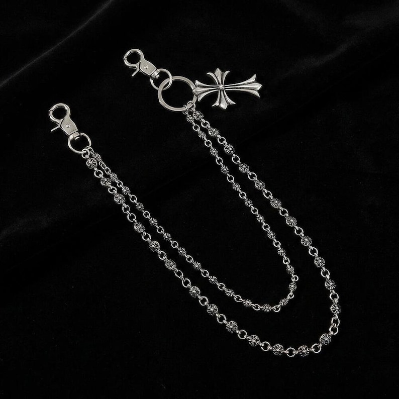 May include: A silver-toned wallet chain with two strands of small, decorative beads. The chain features a cross pendant and two clasps for attachment. The background is a solid black.