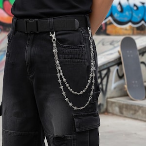 May include: Silver-toned chain accessory with star and bead details, attached to black cargo pants via a belt loop and a pocket. The chain hangs in two strands. A black belt with a buckle is also visible.