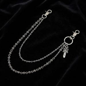 May include: Silver-toned wallet chain with two strands of decorative links. Each strand connects to a clasp at each end. A small dagger charm hangs from a ring on one end. The chain is displayed against a black background.