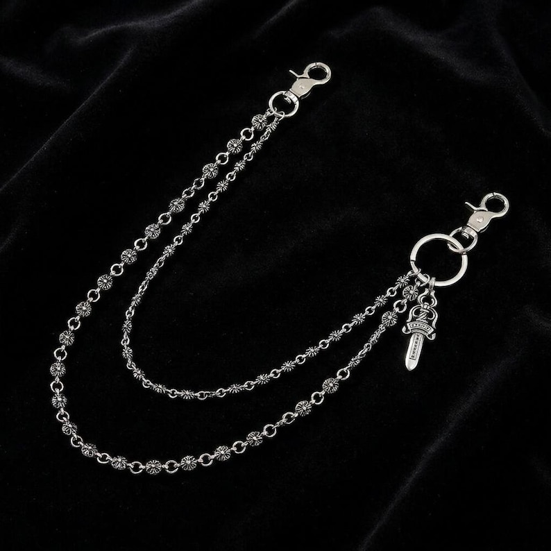 May include: Silver-toned wallet chain with two strands of decorative links. Each strand connects to a clasp at each end. A small dagger charm hangs from a ring on one end. The chain is displayed against a black background.