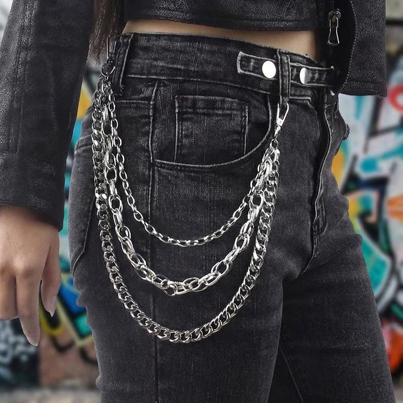 May include: Silver-toned chain accessory with three strands, attached to a belt loop and pocket of black denim jeans. The chains have varying link sizes and hang vertically. The person is wearing a black leather jacket.