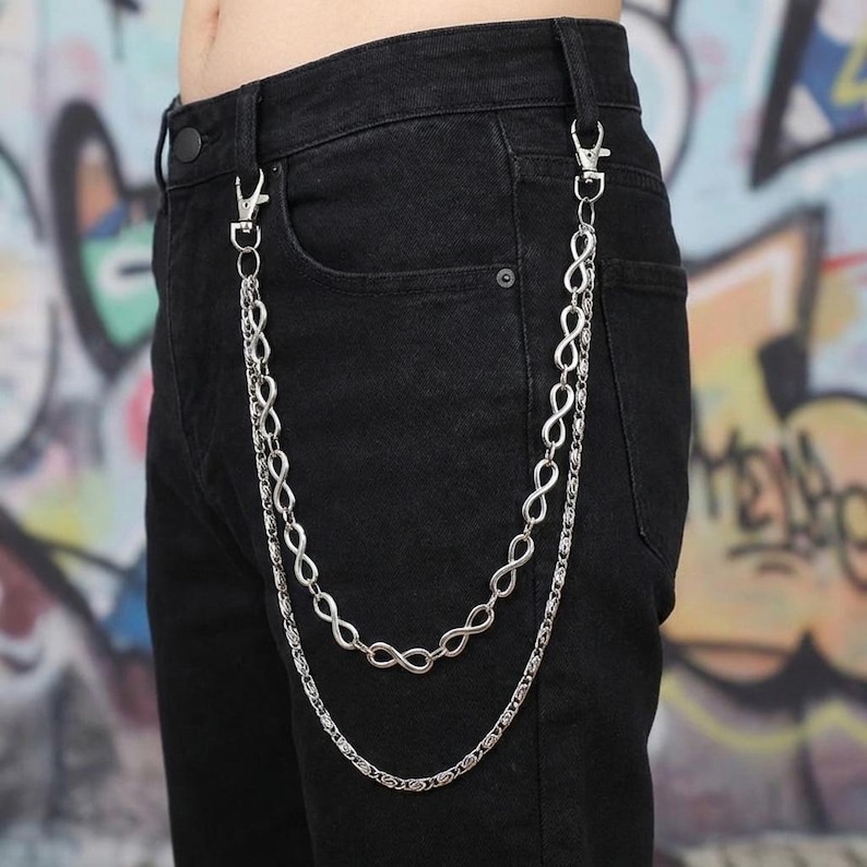 May include: Silver-colored, double-layered chain with infinity symbol links, attached to black denim pants with silver clasps. The chain hangs down the front of the pants. The background has graffiti art.