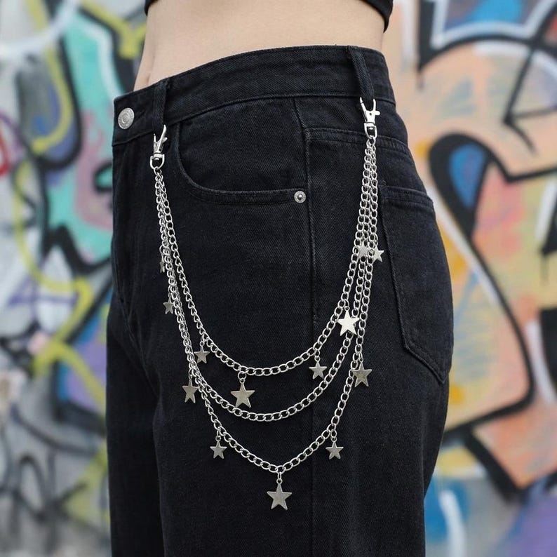 May include: Silver-colored chain belt with star charms. The belt features three layered chains with multiple star-shaped pendants. The belt is attached to the black denim pants with two clasps.