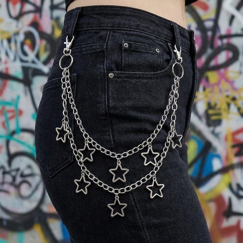 May include: Silver-colored chain belt with star charms. The belt features multiple strands of chain, each adorned with open star-shaped pendants. The belt is attached to the waistband of black denim jeans with a silver clasp.