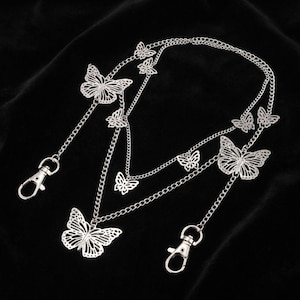 May include: Silver-colored butterfly chain accessory with multiple butterfly pendants and chain links. The accessory has two lobster clasp closures. The butterflies have intricate cut-out designs. The background is black.