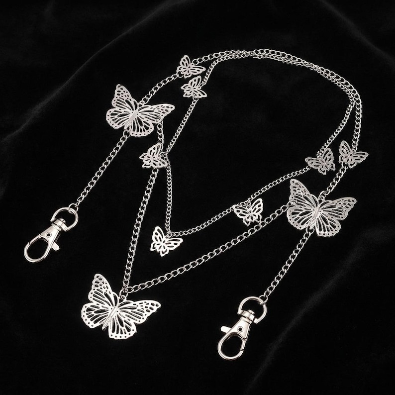 May include: Silver-colored butterfly chain accessory with multiple butterfly pendants and chain links. The accessory has two lobster clasp closures. The butterflies have intricate cut-out designs. The background is black.