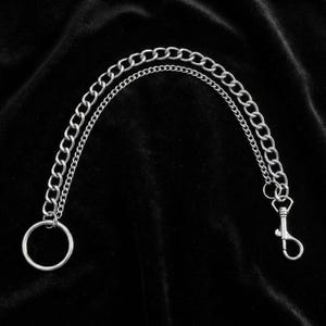 May include: Silver-colored chain with a large ring on one end and a lobster clasp on the other. The chain is composed of two parallel strands of different sizes. The background is a dark, textured fabric.