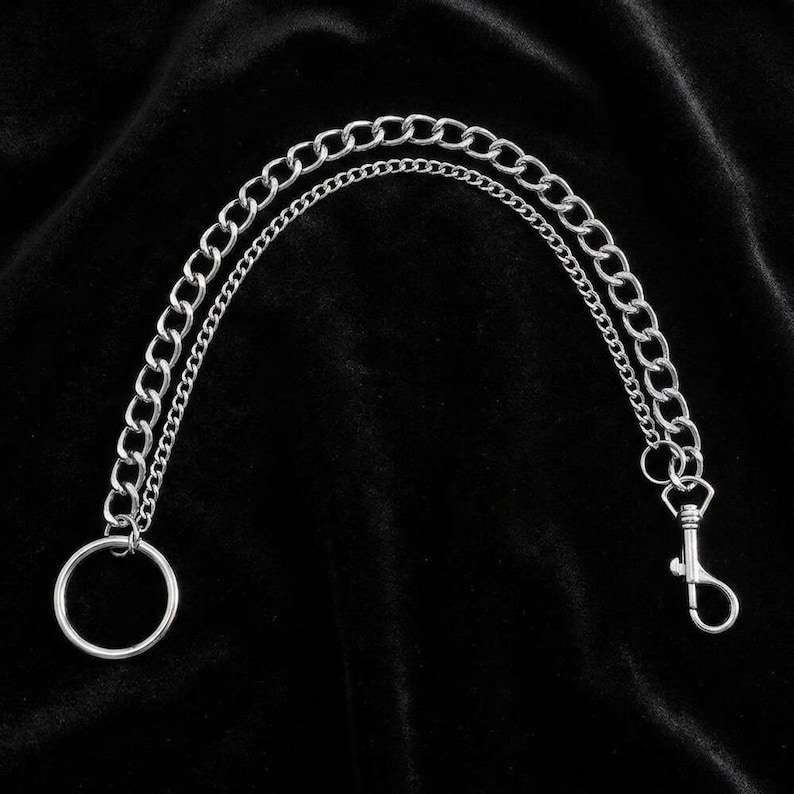 May include: Silver-colored chain with a large ring on one end and a lobster clasp on the other. The chain is composed of two parallel strands of different sizes. The background is a dark, textured fabric.