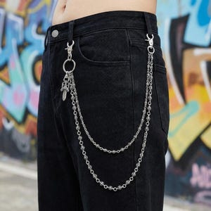 May include: Silver-toned metal wallet chain attached to black denim pants. The chain features two strands of decorative links and a small dagger charm. The pants are worn by a person in front of a graffiti background.