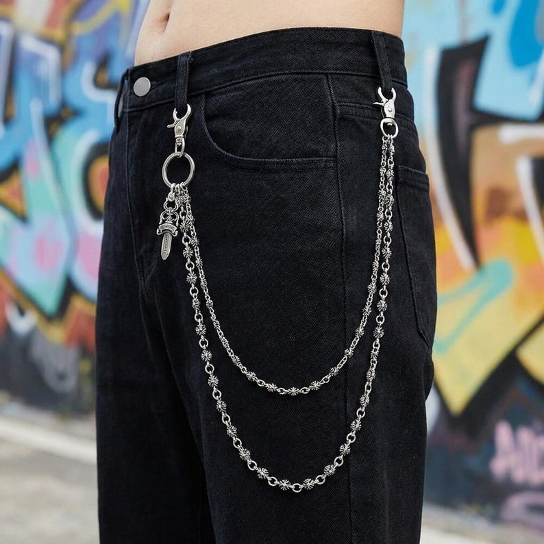 May include: Silver-toned metal wallet chain attached to black denim pants. The chain features two strands of decorative links and a small dagger charm. The pants are worn by a person in front of a graffiti background.